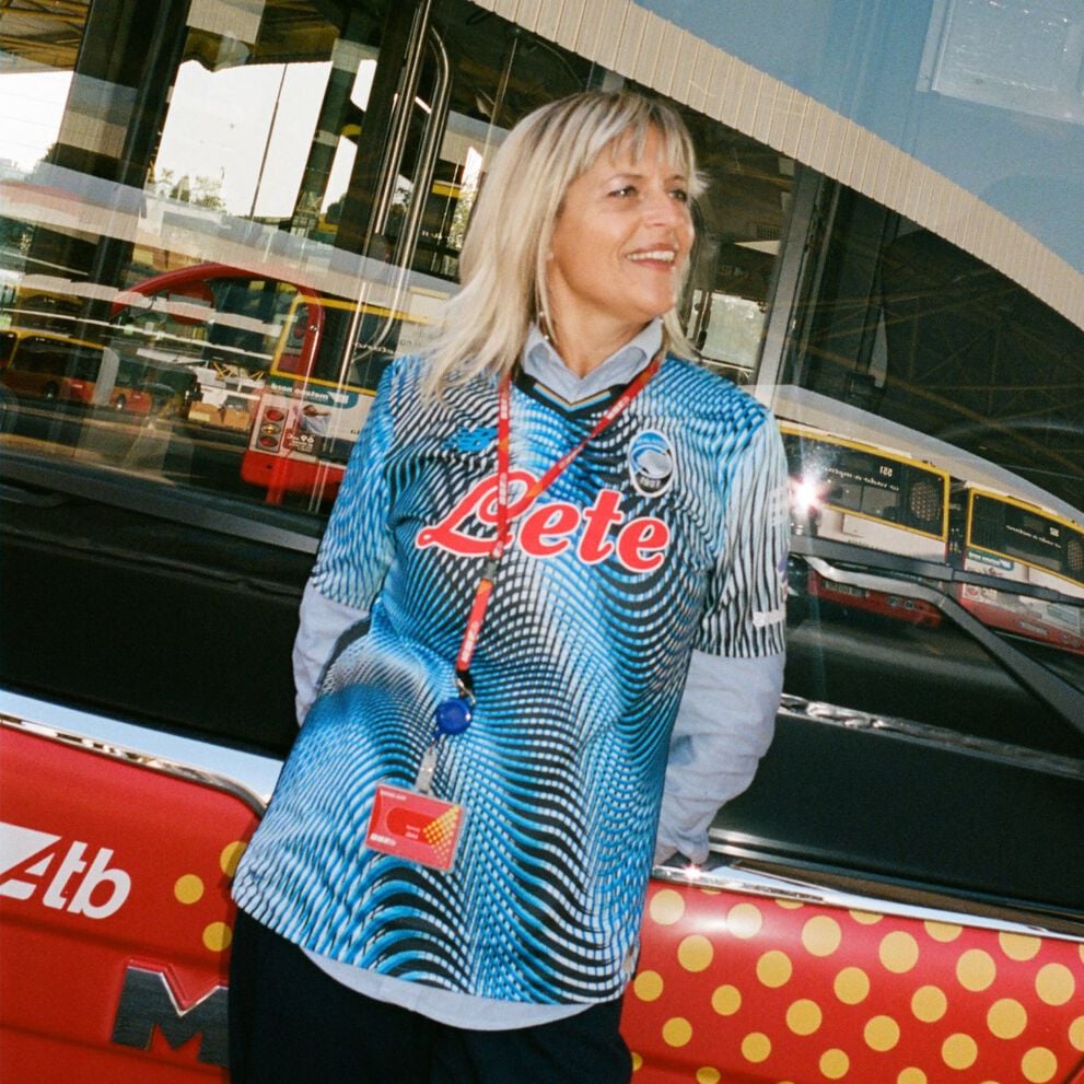 Person wearing a blue and black patterned soccer jersey with the word “Lete” in red across the chest and a team crest near the shoulder, standing in front of a bus with a red and yellow polka-dot design. 